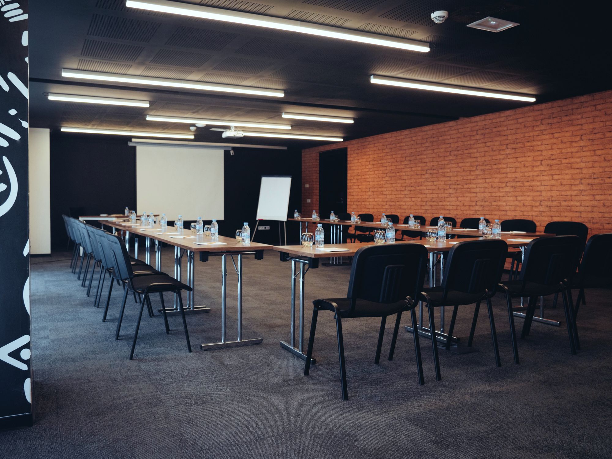 Un salón de conferencias con mesas largas, sillas, botellas de agua y una pantalla de proyector.