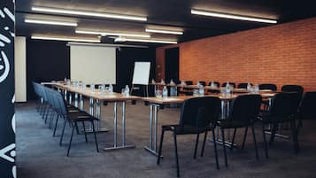 Un salón de conferencias con mesas largas, sillas, botellas de agua y una pantalla de proyector.