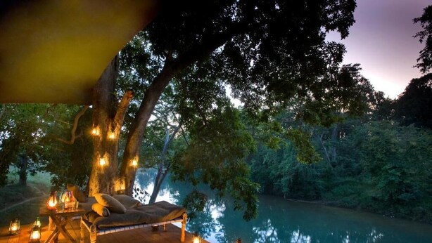 A wooden deck with a bench and lanterns, surrounded by trees and a body of water. - Banjaar Tola A Taj Safari Kanha National Park (Bichhiya)