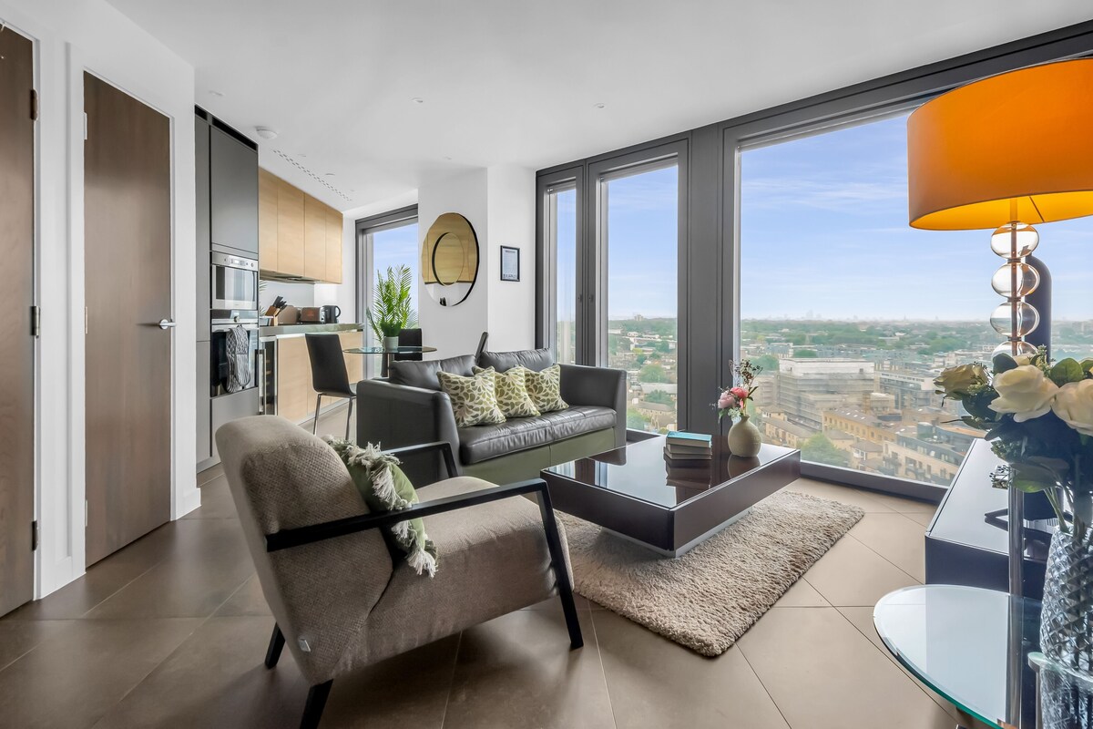 Ein modernes Wohnzimmer mit Sofa, Sessel und Couchtisch, einer Küchenzeile und großen Fenstern mit Blick auf die Stadt.
