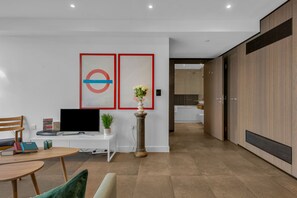 A modern living room with a flat-screen TV, a white desk, a round table, and a green chair. There are two framed artworks on the wall and a potted plant. - Lexicon Apartments (London)
