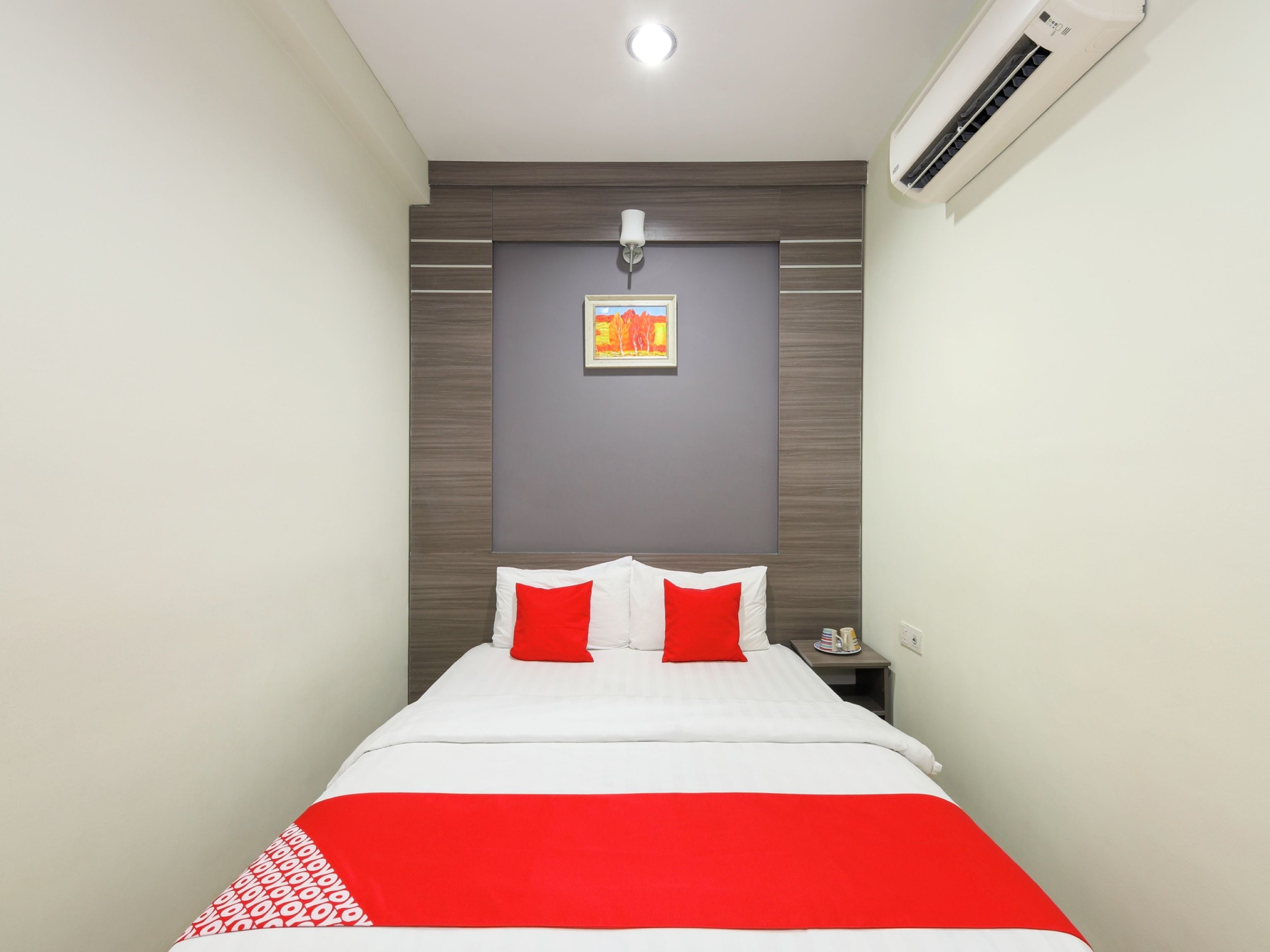 A hotel room with a bed, a nightstand, an air conditioning unit, and a wall-mounted picture.