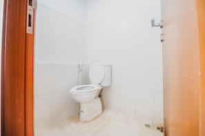 A small, white-tiled bathroom with a white toilet, a wooden door frame, and a visible drain. - Reddoorz Apartment @ Green Lake View Ciputat (Jakarta)