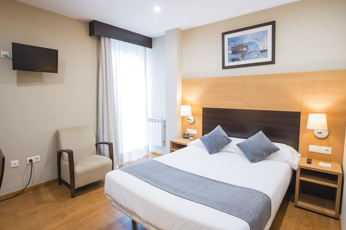 Habitación de hotel con cama, dos lámparas de noche, una silla, televisor y un cuadro en la pared.