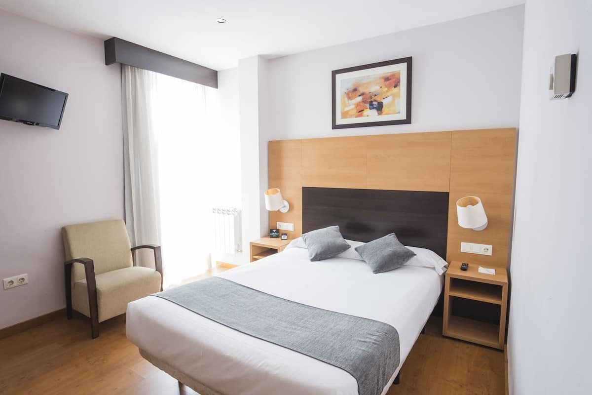 Habitación de hotel con cama, lámparas de noche, una silla, televisor y un cuadro en la pared.