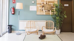 Un vestíbulo con una lámpara moderna, dos sillones de mimbre, una mesita con piñas, un extintor, un radiador y una planta en maceta.
