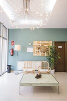 Um hall com um lustre moderno, duas cadeiras de vime, uma pequena mesa com pinhas, um extintor de incêndio, um radiador e uma planta em vaso.