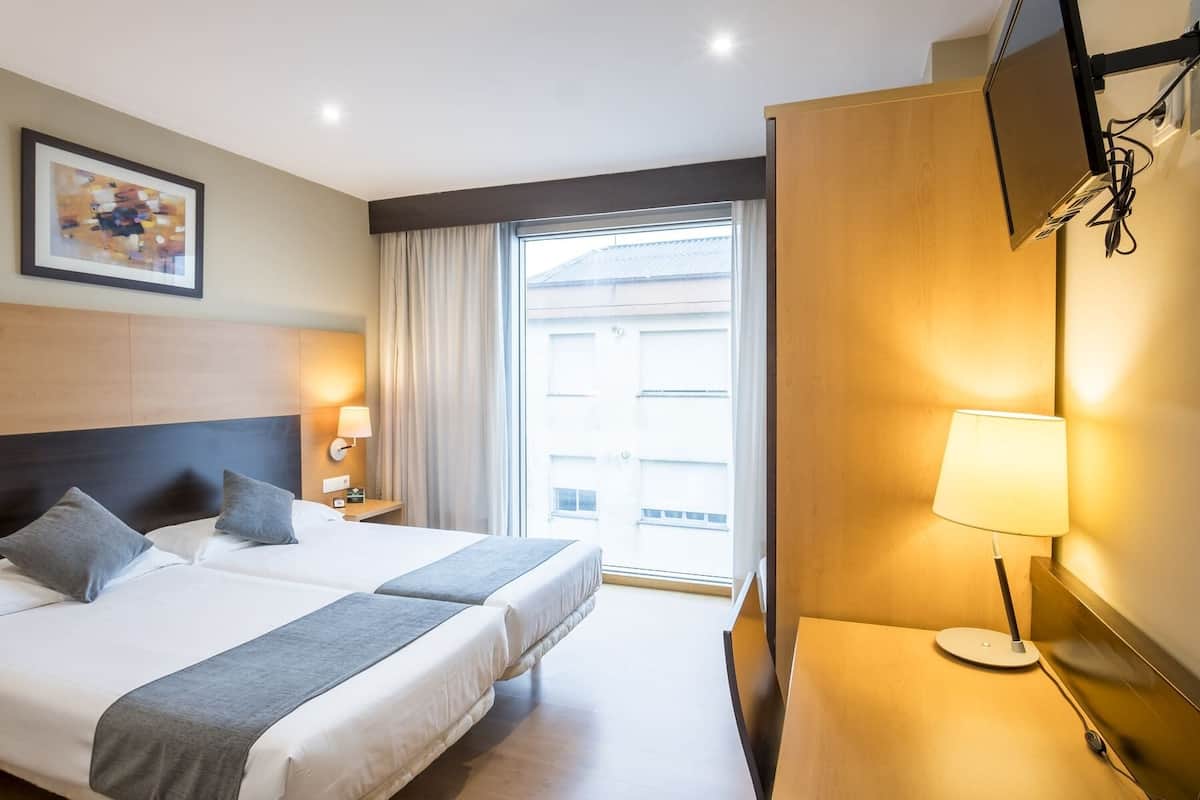 Una habitación de hotel con cama, lámparas de noche, televisión y ventana con cortinas.