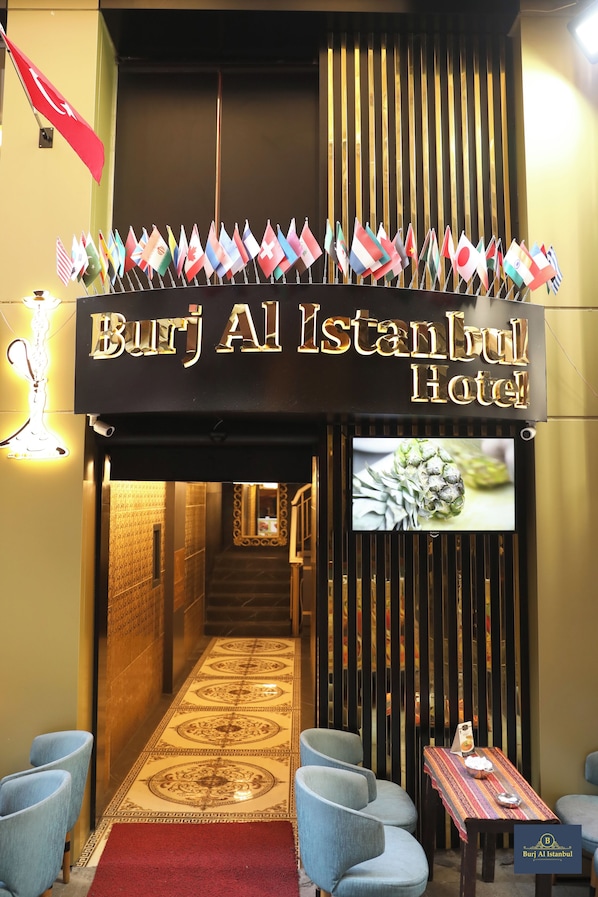 Der Eingang des Burj Al Istanbul Hotels mit Schildern, Flaggen, einem Bildschirm und einer Sitzgelegenheit.