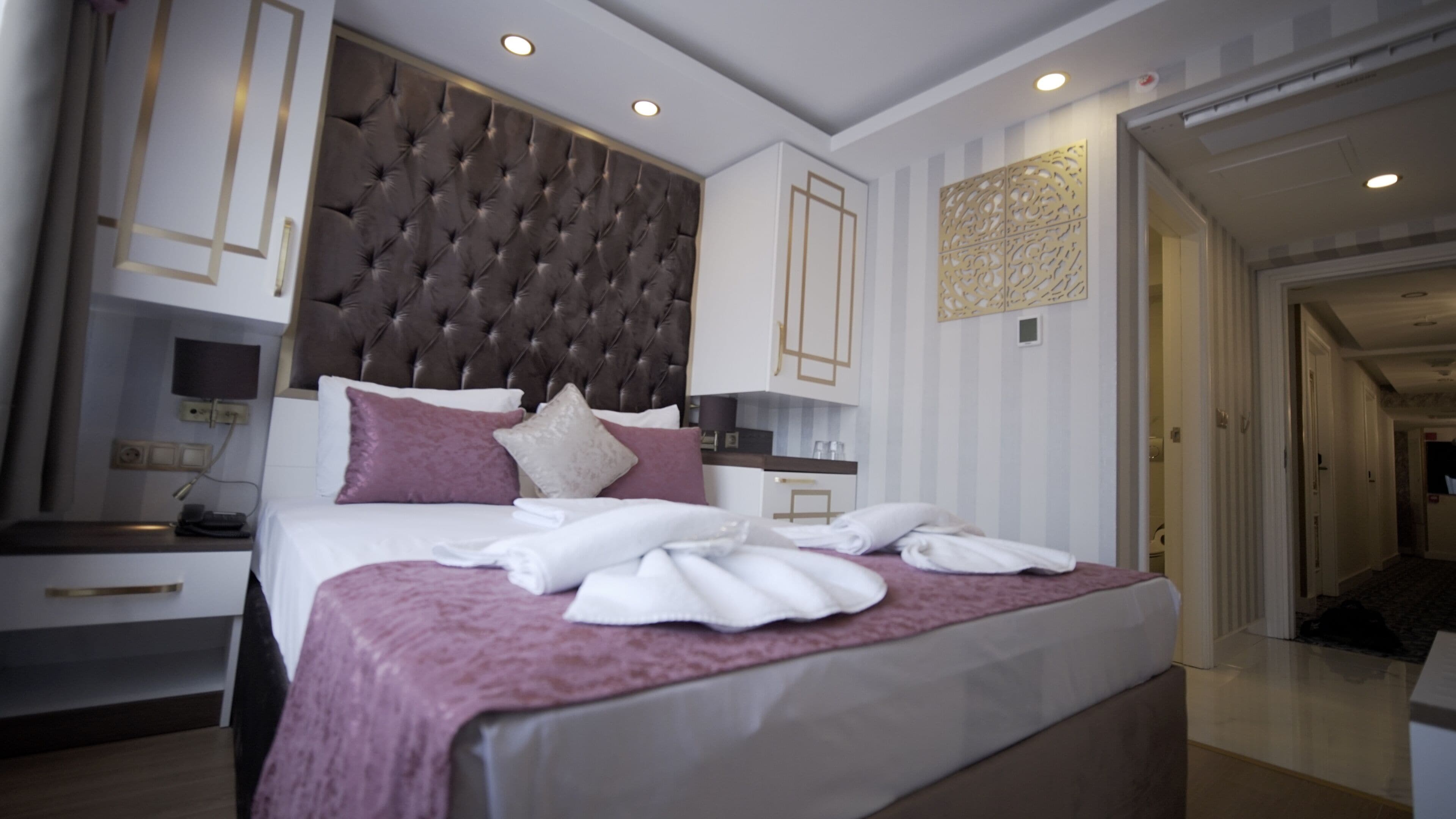 A modern hotel room with a large bed, a built-in headboard, a bedside table, and a small desk with a chair.