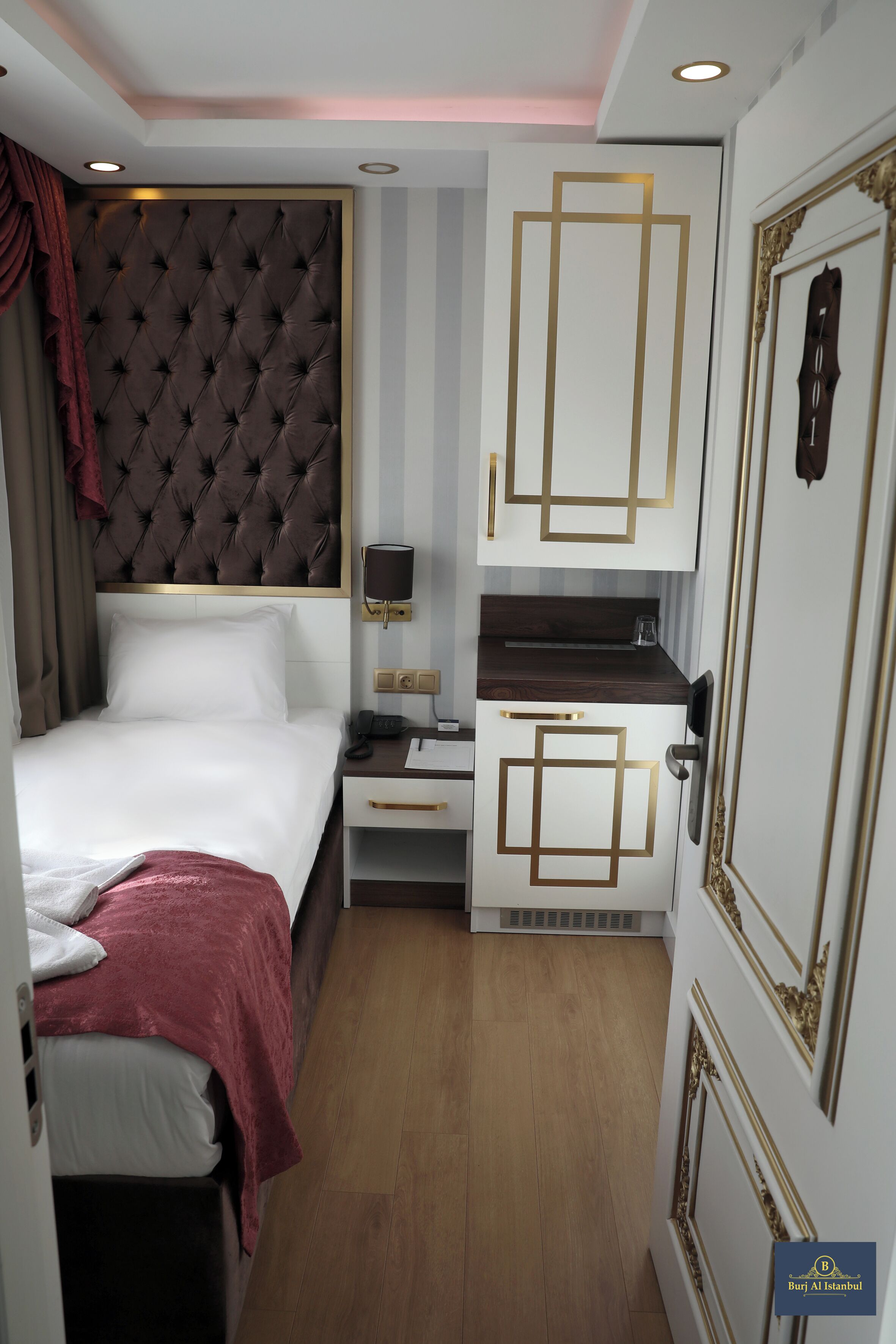 A compact hotel room with a tufted headboard, a bed with white linens, a small desk with a lamp, and a kitchenette with decorative gold accents.