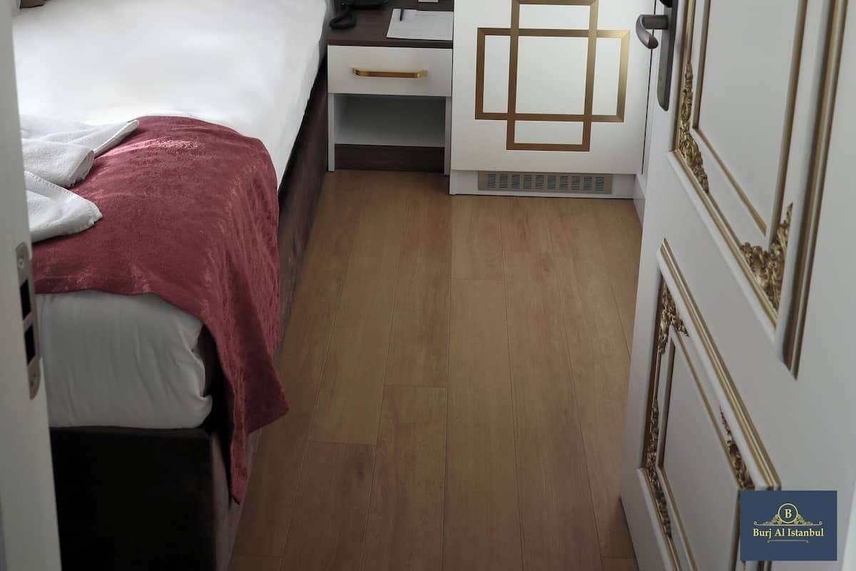 Ein kompaktes Hotelzimmer mit einer gepolsterten Kopfstütze, einem Bett mit weißen Bettwäsche, einem kleinen Schreibtisch mit Lampe und einer Küchenzeile mit goldenen Akzenten.
