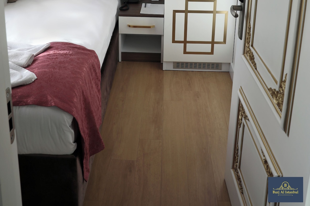 Une chambre d’hôtel compacte dotée d’une tête de lit capitonnée, d’un lit avec du linge de lit blanc, d’un petit bureau avec une lampe et d’une kitchenette agrémentée de touches décoratives dorées.