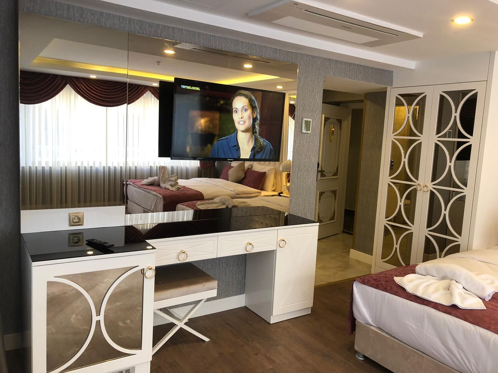 A hotel room with a large flat-screen TV mounted on the wall, a desk with drawers, a bed with a red headboard, and a decorative mirror.