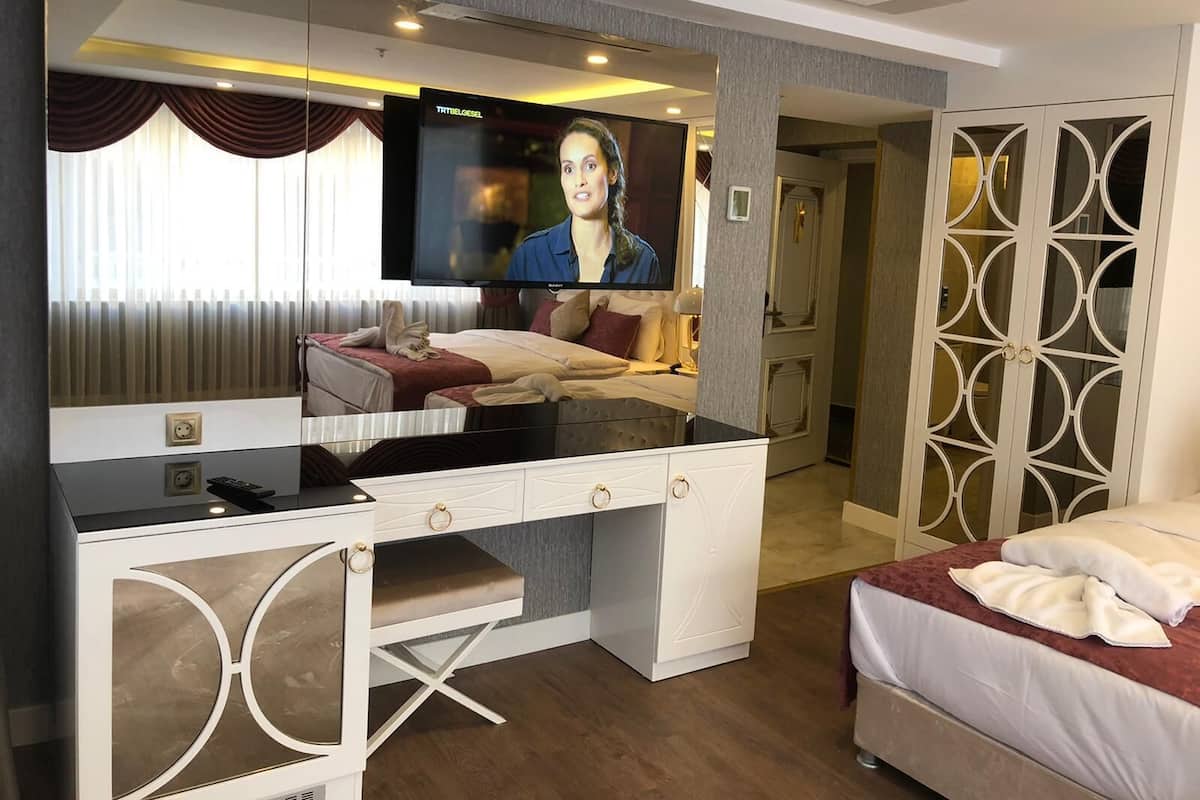 Ein Hotelzimmer mit einem großen Flachbildfernseher an der Wand, einem Schreibtisch mit Schubladen, einem Bett mit rotem Kopfteil und einem dekorativen Spiegel.