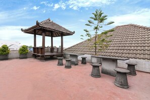 A traditional pavilion with a tiled roof, stone benches, and a tree. - Hotel Padjadjaran 1 (Depok)