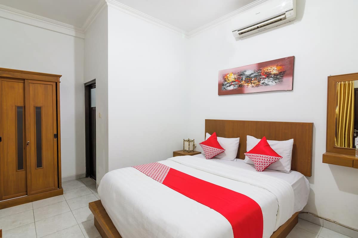 A neatly arranged bedroom with a large bed, a wooden wardrobe, a nightstand with a lamp, and a painting on the wall.