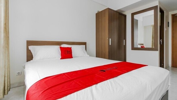 A hotel room with a bed, a red throw blanket, white bedding, and a wooden wardrobe. - Reddoorz Apartment @ Margonda Residence Tower 2 (Depok)