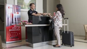 A reception desk with a man and a woman exchanging a card, a red sign with hotel services, and a suitcase. - Reddoorz Apartment @ Margonda Residence Tower 2 (Depok)