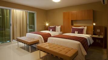 A hotel room with two beds, a bench, a wooden headboard, and a sliding glass door.