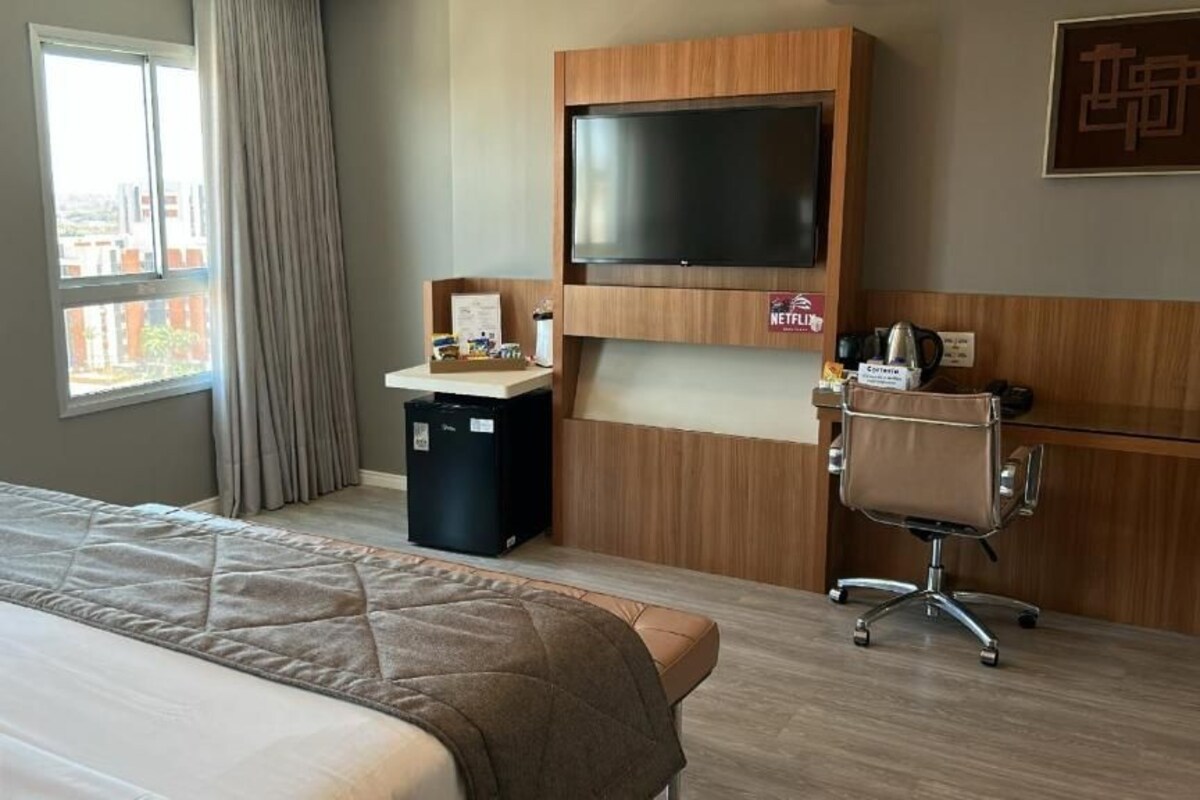 A hotel room with a bed, a desk with a flat-screen TV, a chair, and a small kitchenette area.