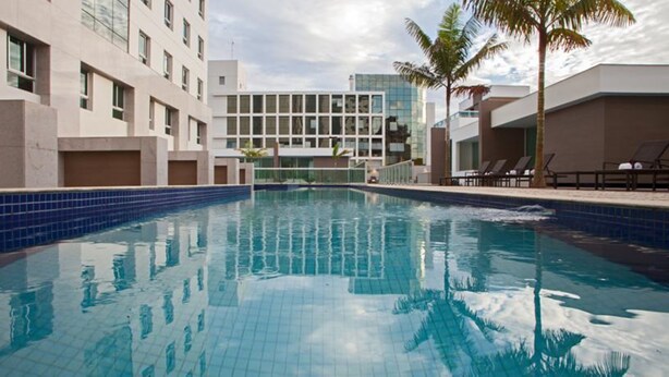 A swimming pool with clear blue water, surrounded by modern buildings and palm trees. - Jade Hotel (Guará)