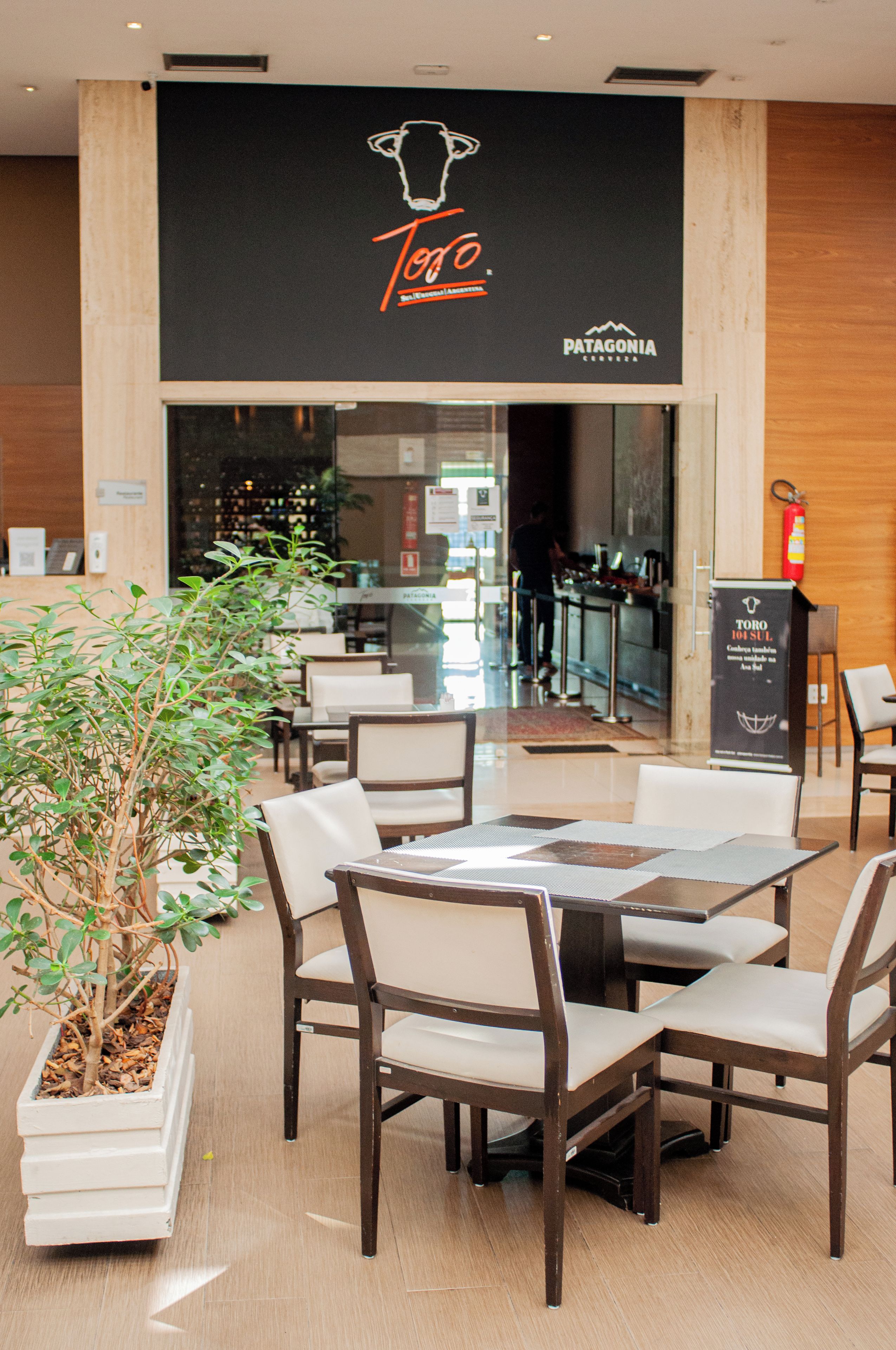 A restaurant with a black sign featuring a cow logo and the name 'Toro.' There are wooden chairs and tables, a potted plant, and a fire extinguisher on the wall.