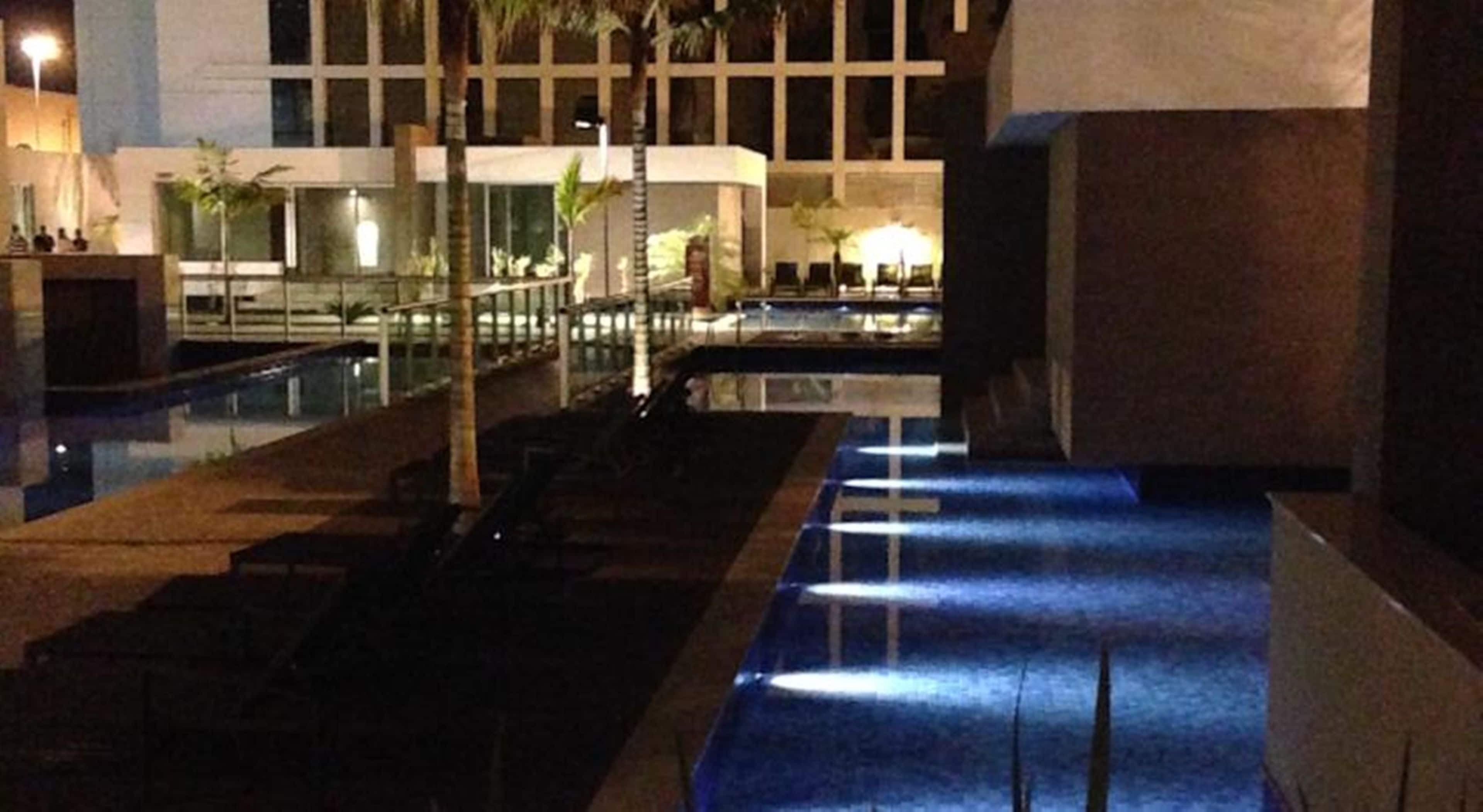 A nighttime view of a hotel pool area with a water feature, palm trees, and a building in the background.