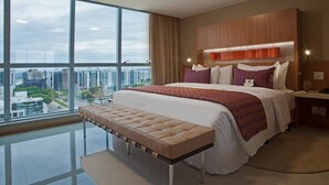 A modern hotel room with a large bed, a bench, a nightstand, and a view of the city. - Jade Hotel (Guará)