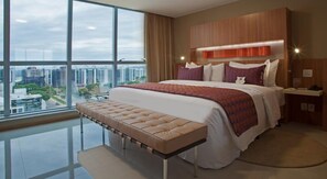 Una habitación de hotel moderna con una cama grande, un banco, una mesita de noche y vistas a la ciudad.