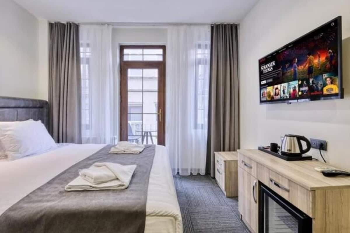 A hotel room with a bed, a TV mounted on the wall, a desk with a kettle, and a window with curtains.
