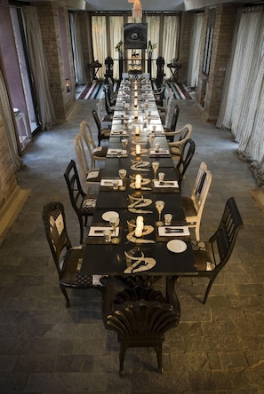 A long dining table set for a meal, flanked by chairs, with a brick wall and curtains in the background.