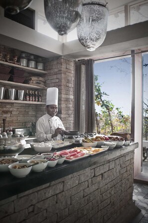 A chef in a kitchen with a brick wall, a large window, and various food items displayed on the counter. - Pashan Garh A Taj Safari Panna National Park (Pawai)
