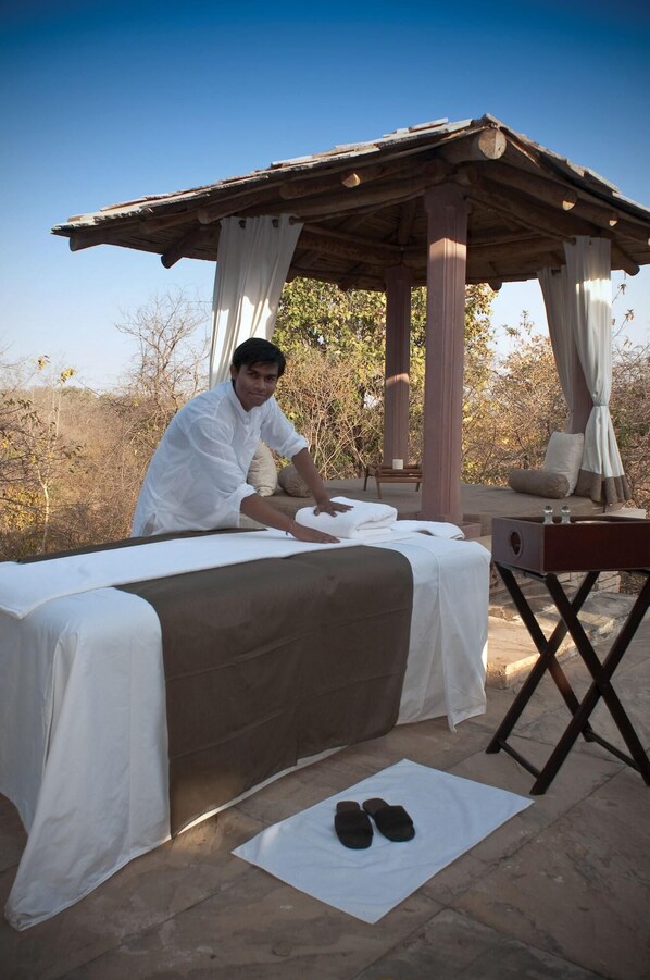 A person preparing a bed with a white sheet and a brown blanket in an outdoor setting with a wooden structure and trees in the background.
