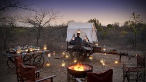 A rustic outdoor dining setup with a fire pit, wooden chairs, a covered wagon with a person inside, and lanterns.