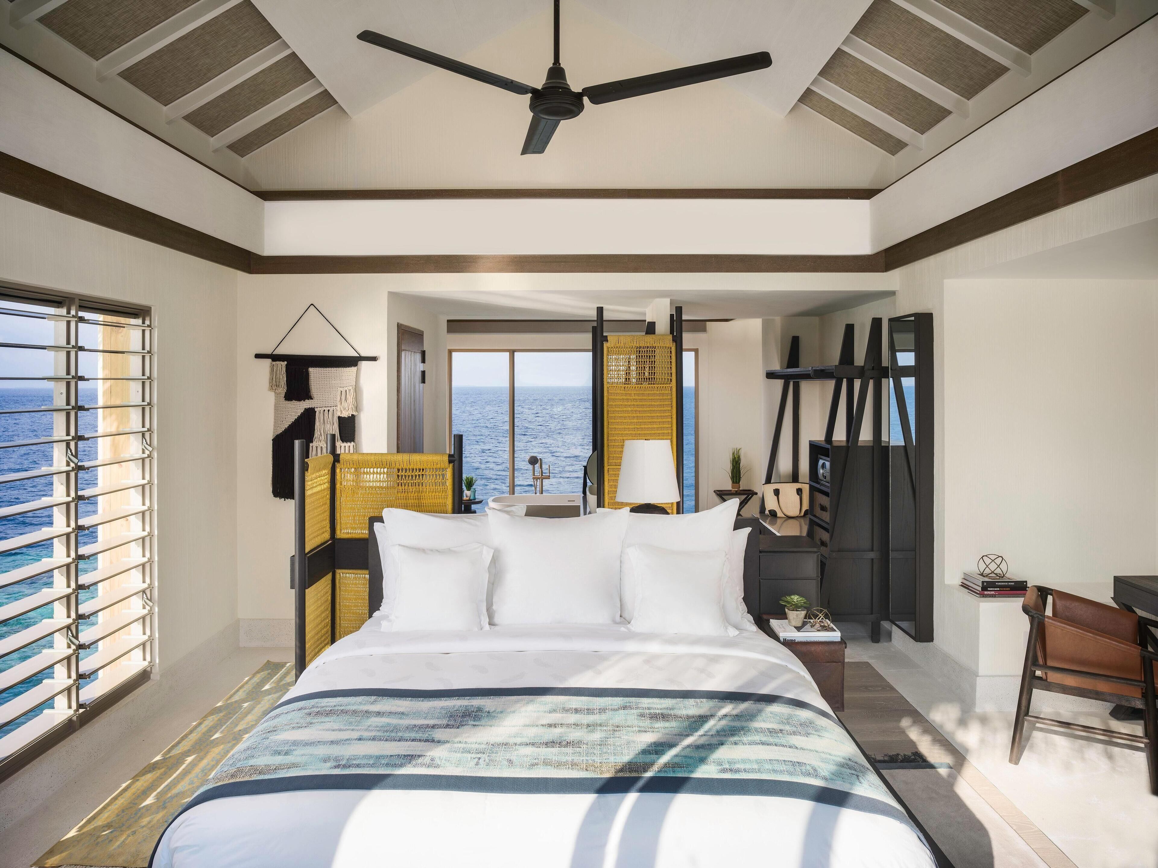 Une chambre d’hôtel dotée d’un grand lit, d’un ventilateur de plafond, d’une vue sur l’océan et d’un coin salon avec une table et des chaises.