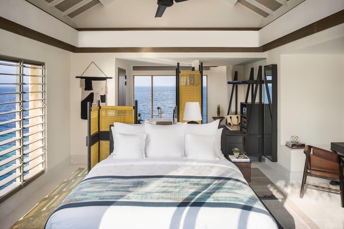 Quarto de hotel com uma cama grande, ventoinha no teto, vista para o mar e um espaço de estar com mesa e cadeiras.