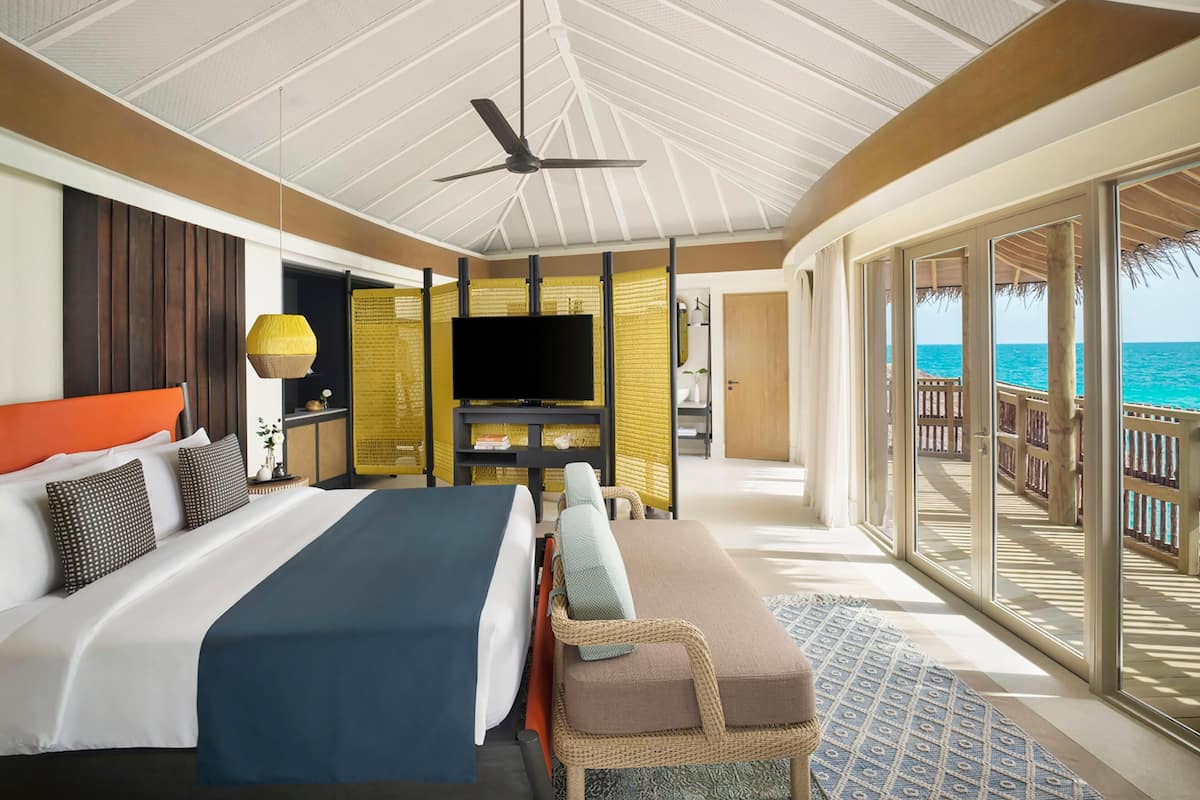 Quarto de hotel com cama, televisão, varanda e vista para o mar, e ventoinha no teto.