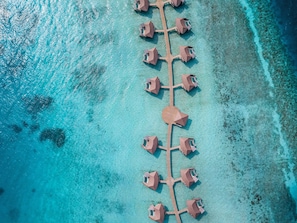 Vista aerea di un resort con un edificio centrale e diverse bungalow sull'acqua collegati da una passerella in legno.