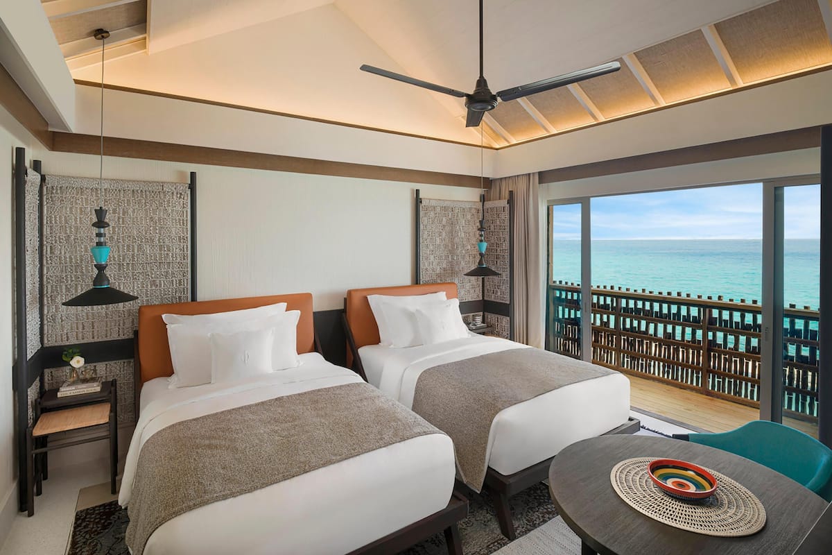 Quarto de hotel com duas camas, um ventilador de teto, vista para o oceano e varanda com grade de madeira.