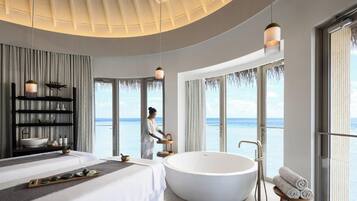 A modern hotel room with a large bathtub, a bed, a vanity with a bowl sink, and a view of the ocean.