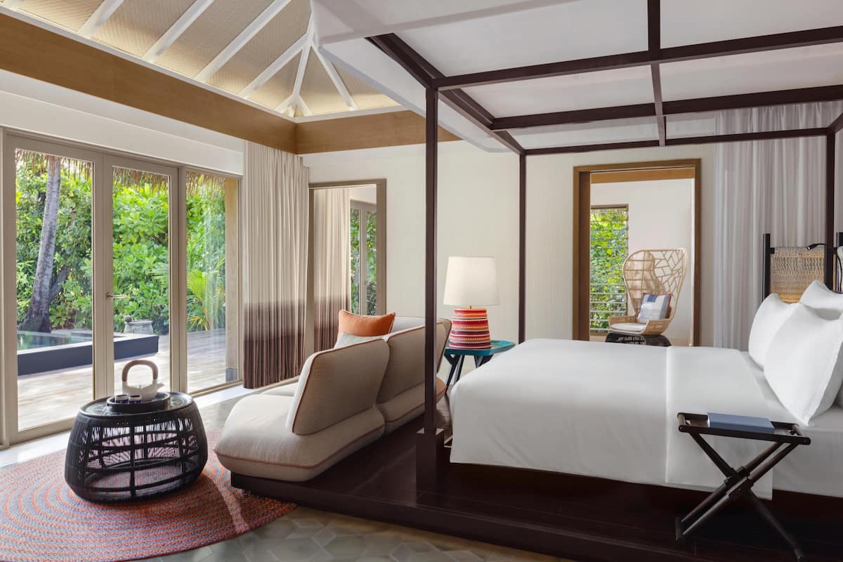 Quarto de hotel com cama, sofá, luminária, mesa pequena e vista para área verde através de grandes janelas.
