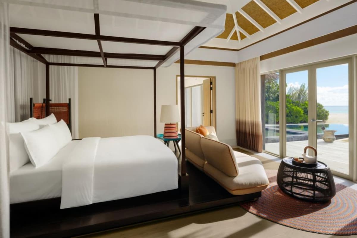Quarto de hotel com cama, sofá, mesinha com abajur, objeto decorativo e vista para o exterior.