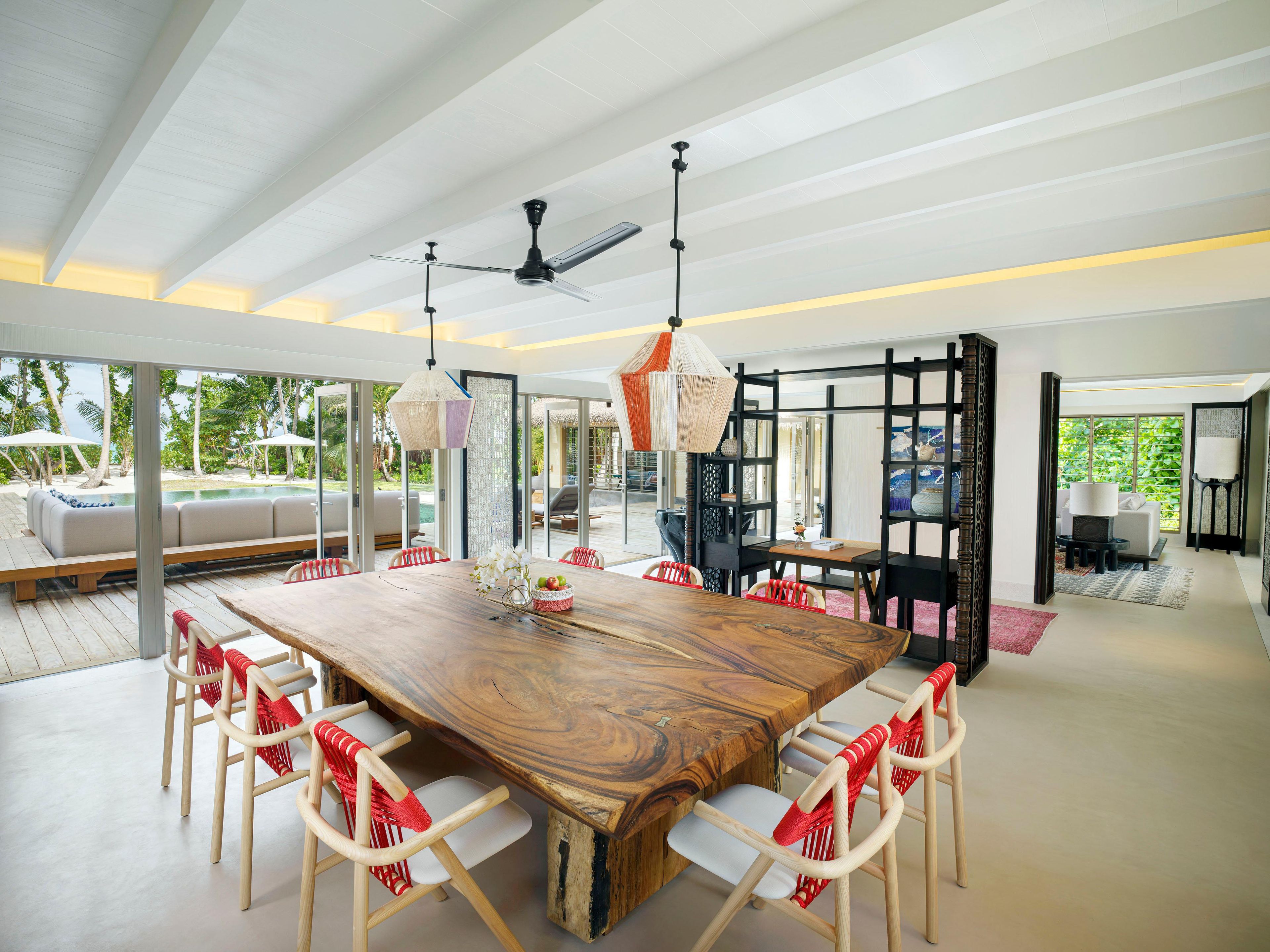 A spacious dining area with a large wooden table, white chairs with red accents, and a ceiling fan.