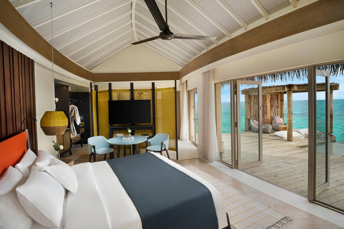 Quarto com cama, ventoinha no teto, televisão, área de estar com cadeiras e vista para o mar através de grandes portas de vidro.