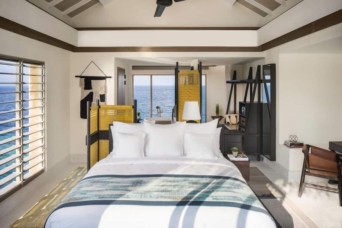 Quarto de hotel com cama, ventoinha no teto, vista para o mar e secretária com cadeira.