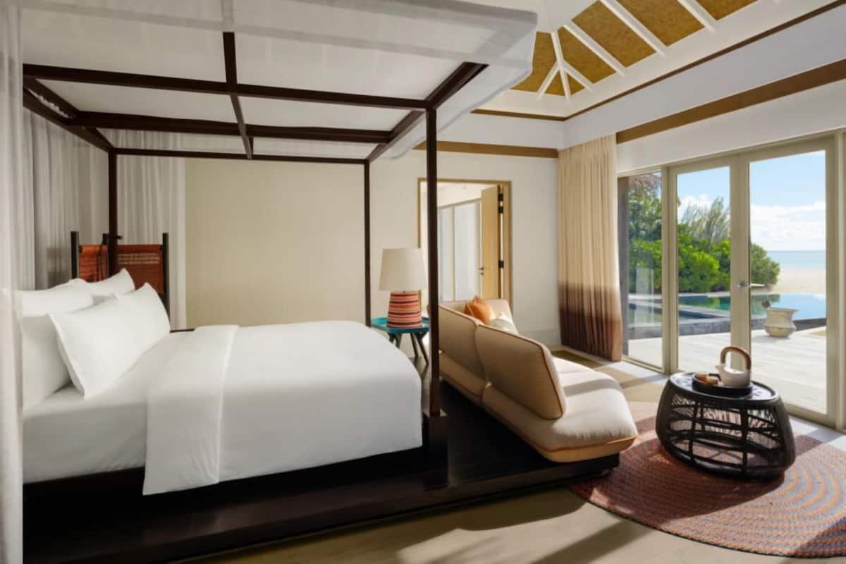 Quarto de hotel com cama, sofá, mesinha com abajur, objeto decorativo e vista para o exterior.