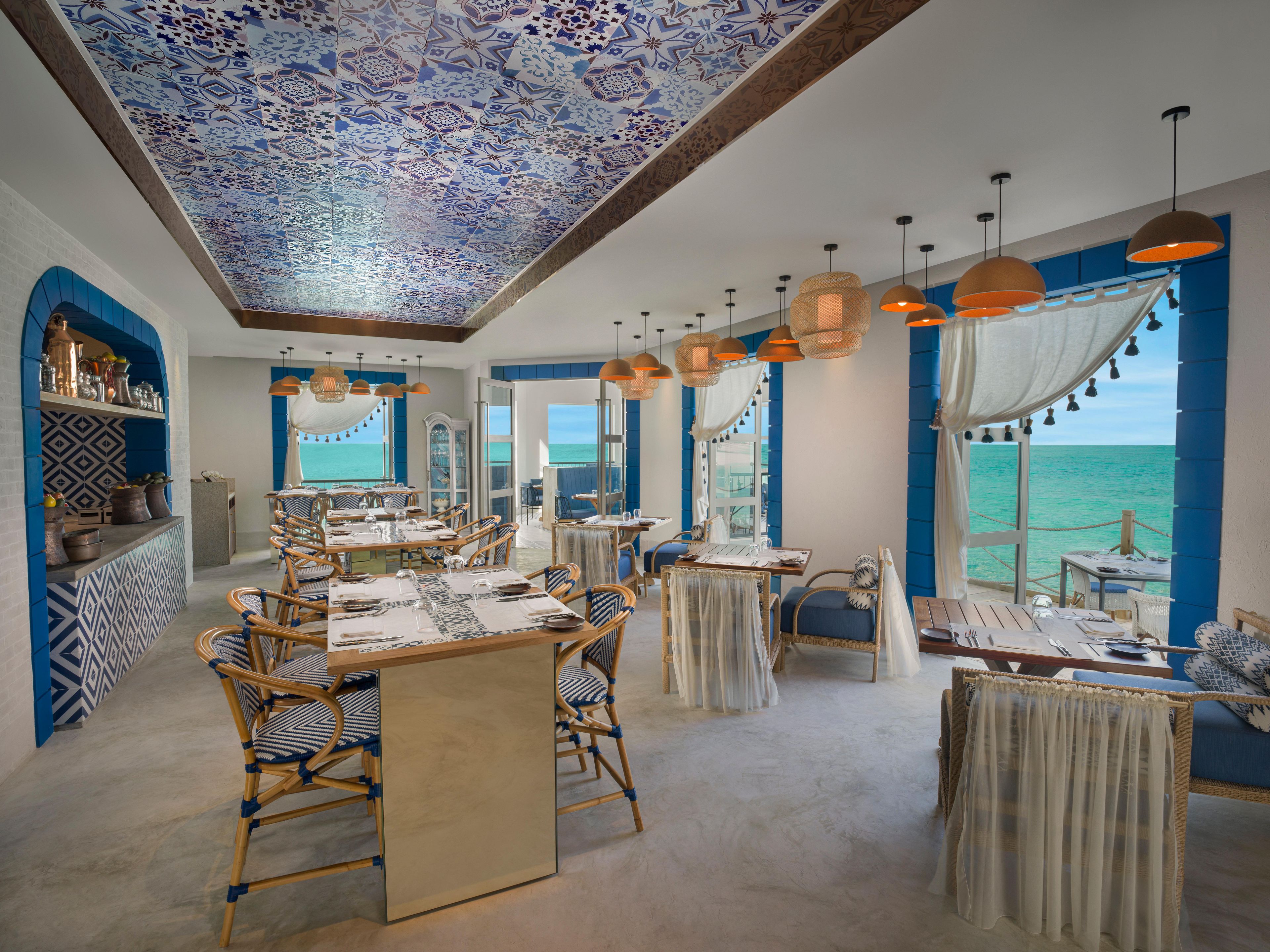 A restaurant with a view of the sea, featuring blue and white patterned walls, wicker furniture, and hanging pendant lights.