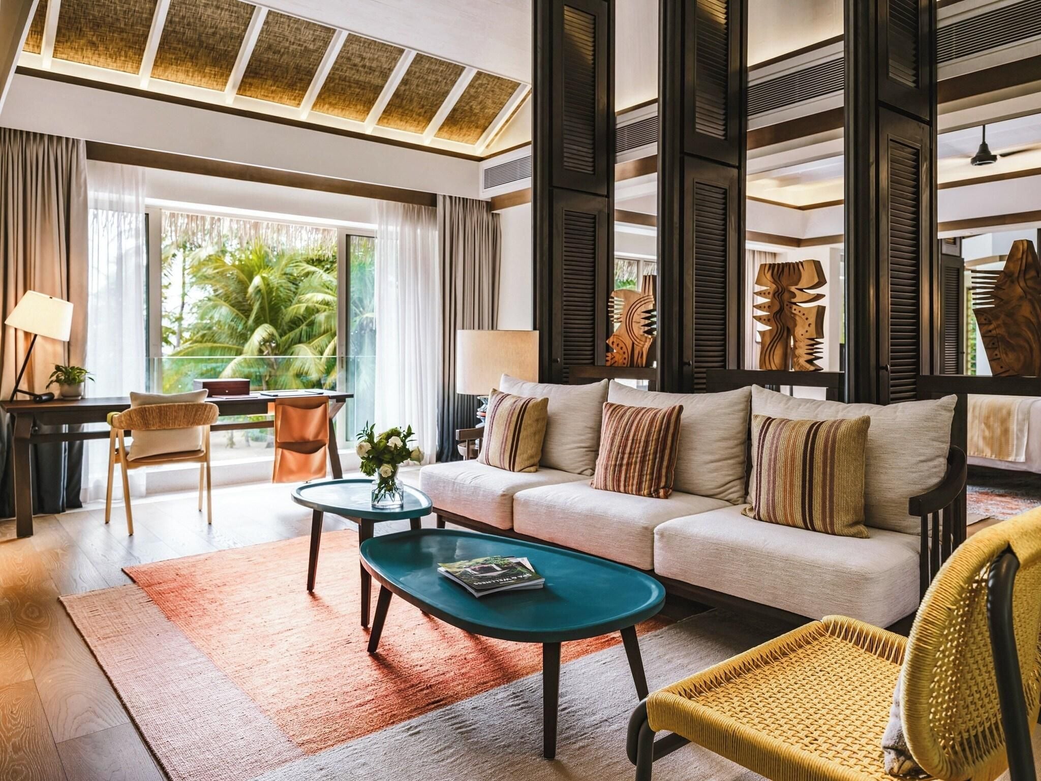 A modern living room with a sofa, coffee table, and chairs, large windows with a view of greenery, and a ceiling with a geometric pattern.