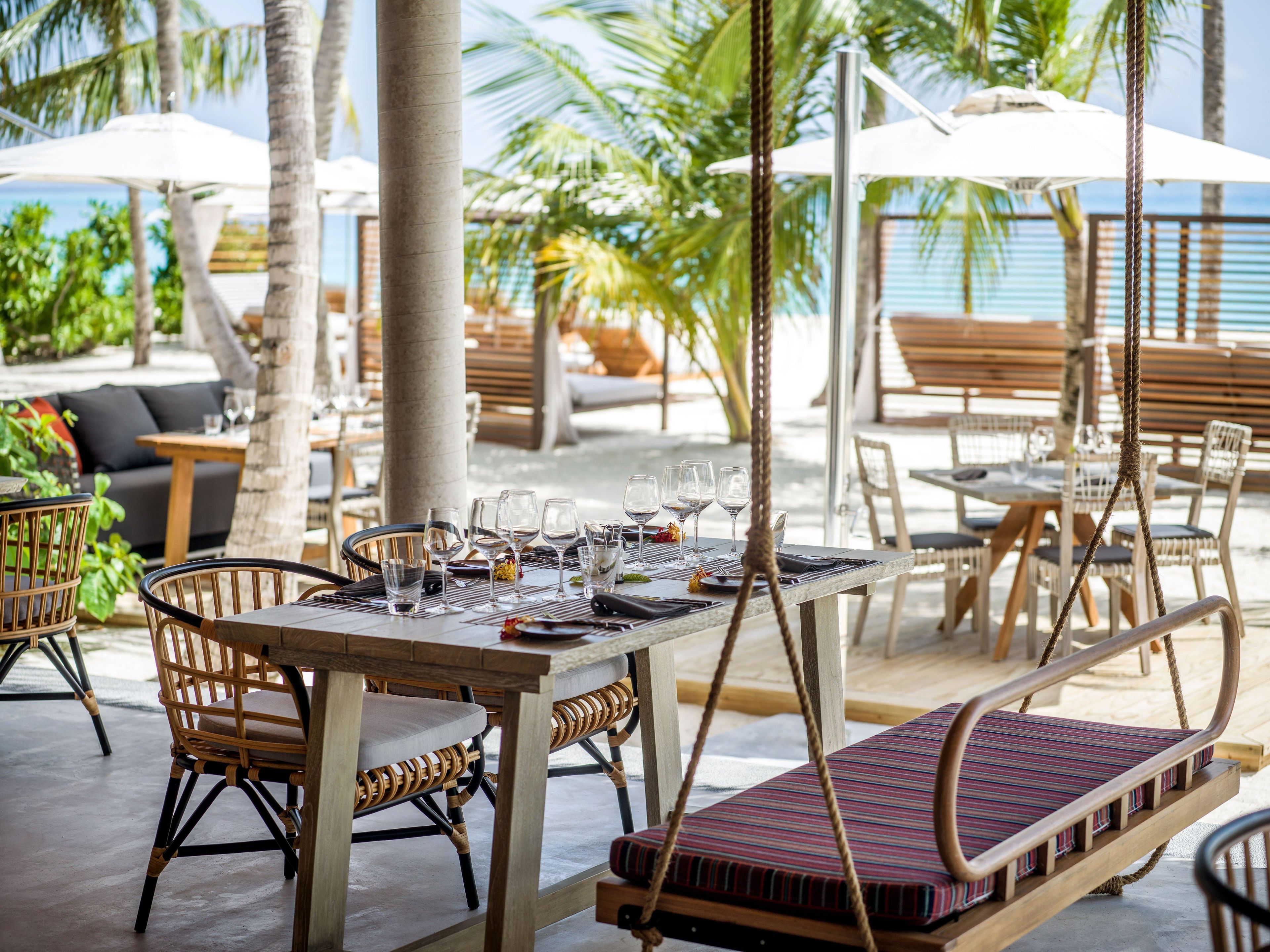An outdoor dining area with tables, chairs, and umbrellas, surrounded by palm trees and a view of the ocean.